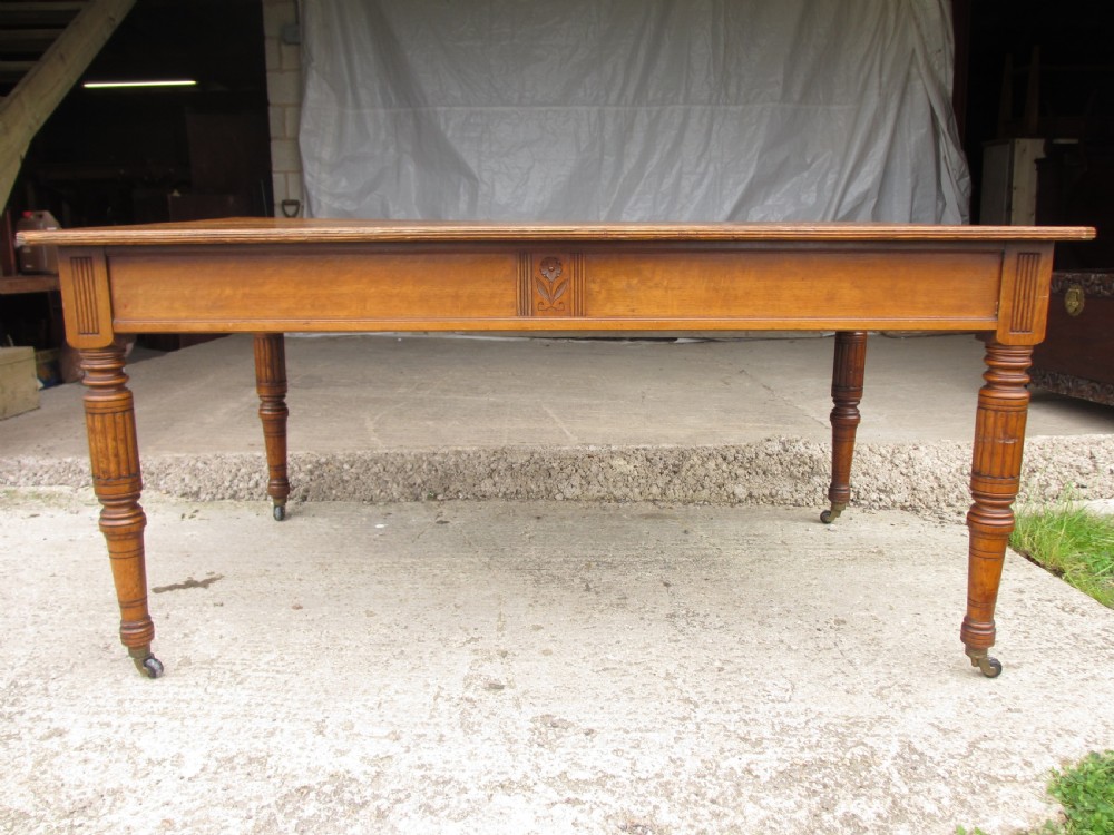 An Edwardian Arts and Crafts warm brown oak library table featuring two front drawers, a carved central floral panel, turned legs, and brass handles on castors. View XII