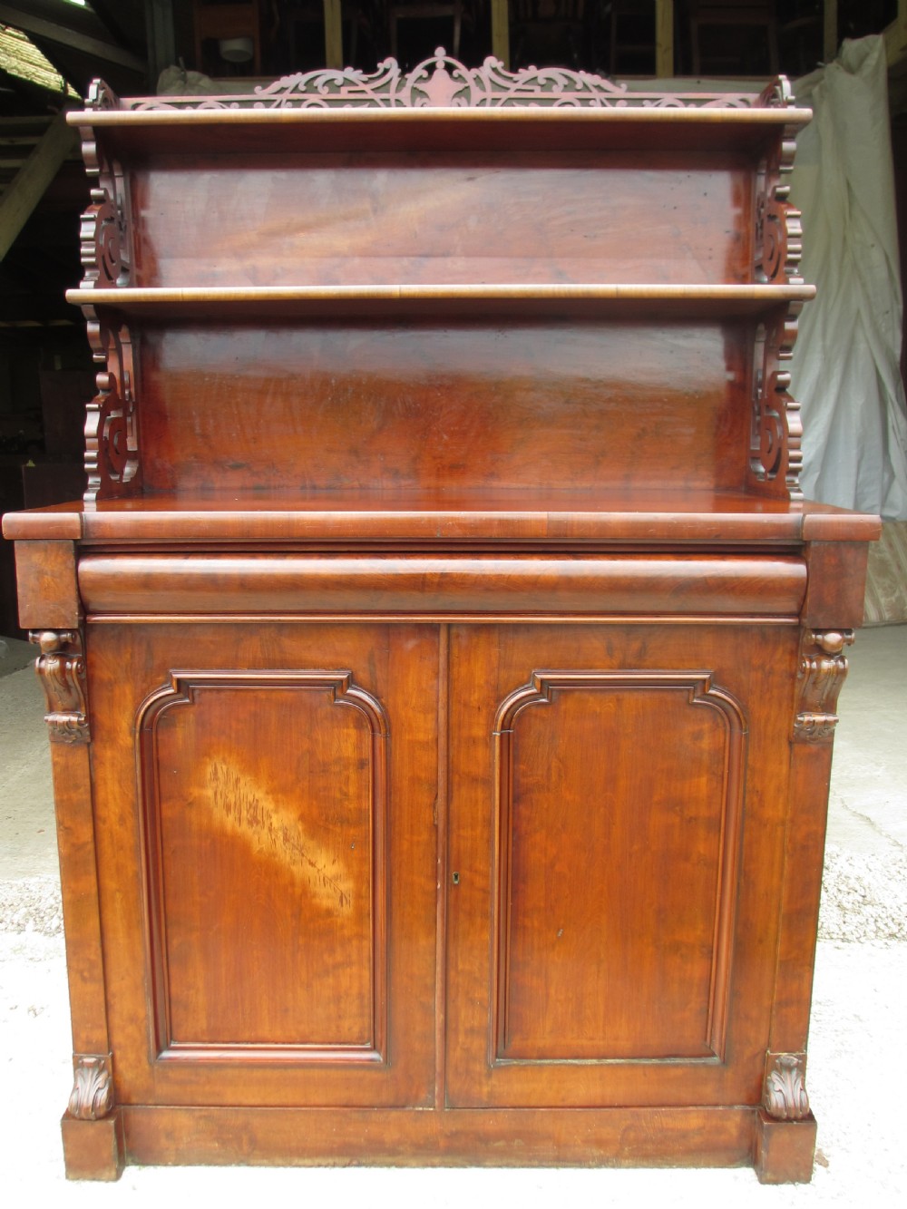 A Victorian mahogany chiffonier featuring two lower cupboard doors with figured plum pudding veneer panels, topped with two open display shelves supported by carved scroll brackets and a fretwork cresting.