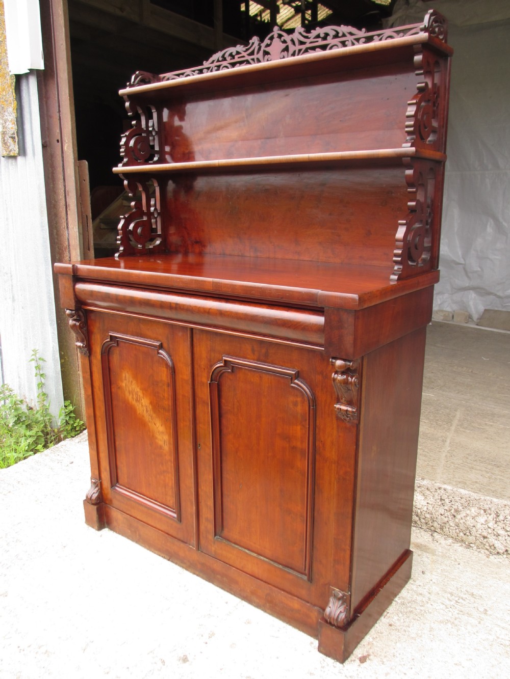 A Victorian mahogany chiffonier featuring two lower cupboard doors with figured plum pudding veneer panels, topped with two open display shelves supported by carved scroll brackets and a fretwork cresting. View II