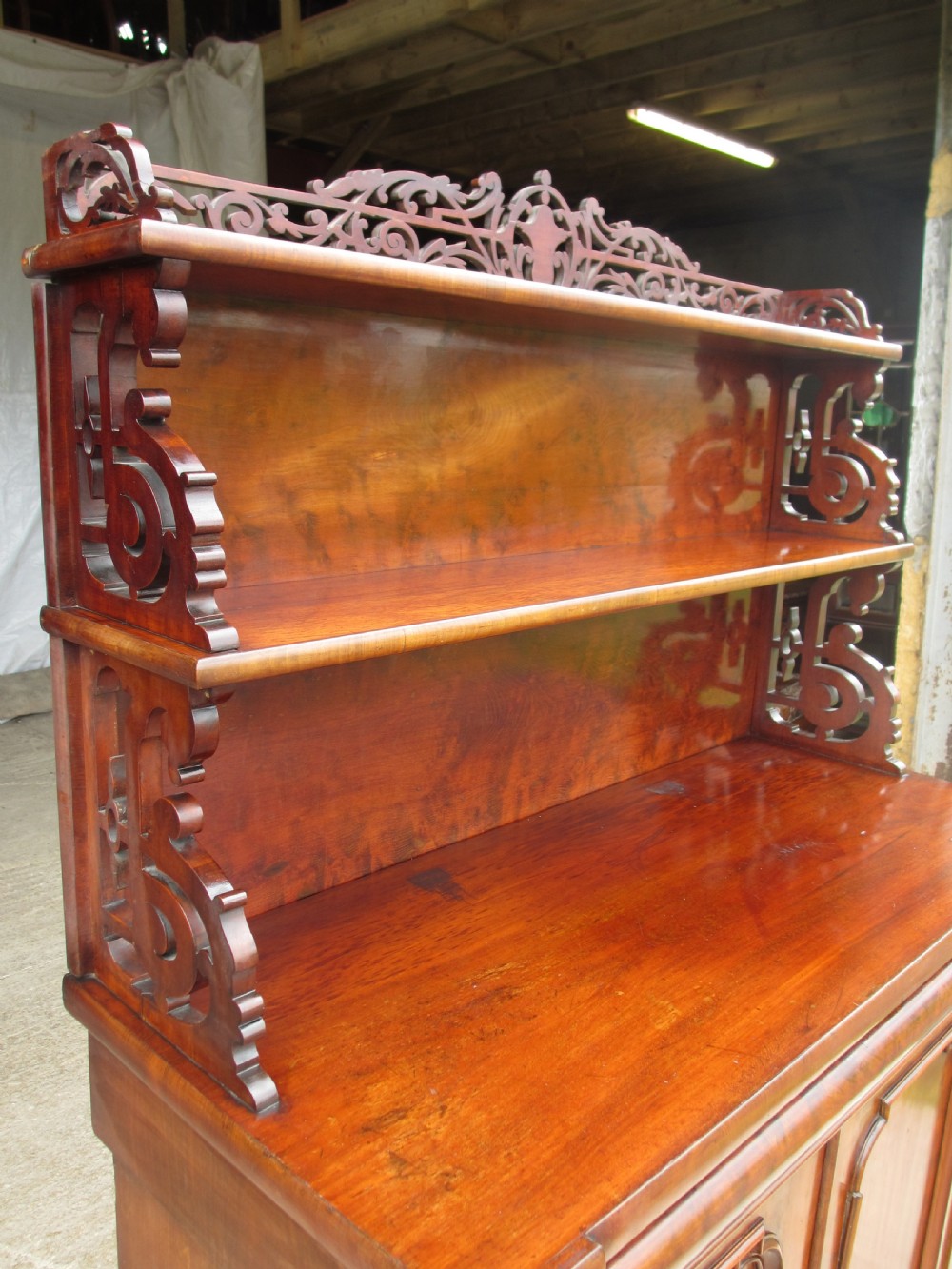 A Victorian mahogany chiffonier featuring two lower cupboard doors with figured plum pudding veneer panels, topped with two open display shelves supported by carved scroll brackets and a fretwork cresting. View III