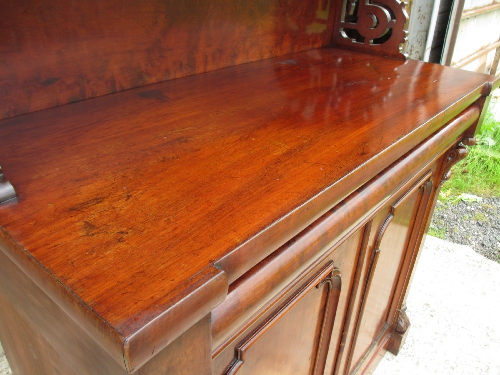 A Victorian mahogany chiffonier featuring two lower cupboard doors with figured plum pudding veneer panels, topped with two open display shelves supported by carved scroll brackets and a fretwork cresting. View VI