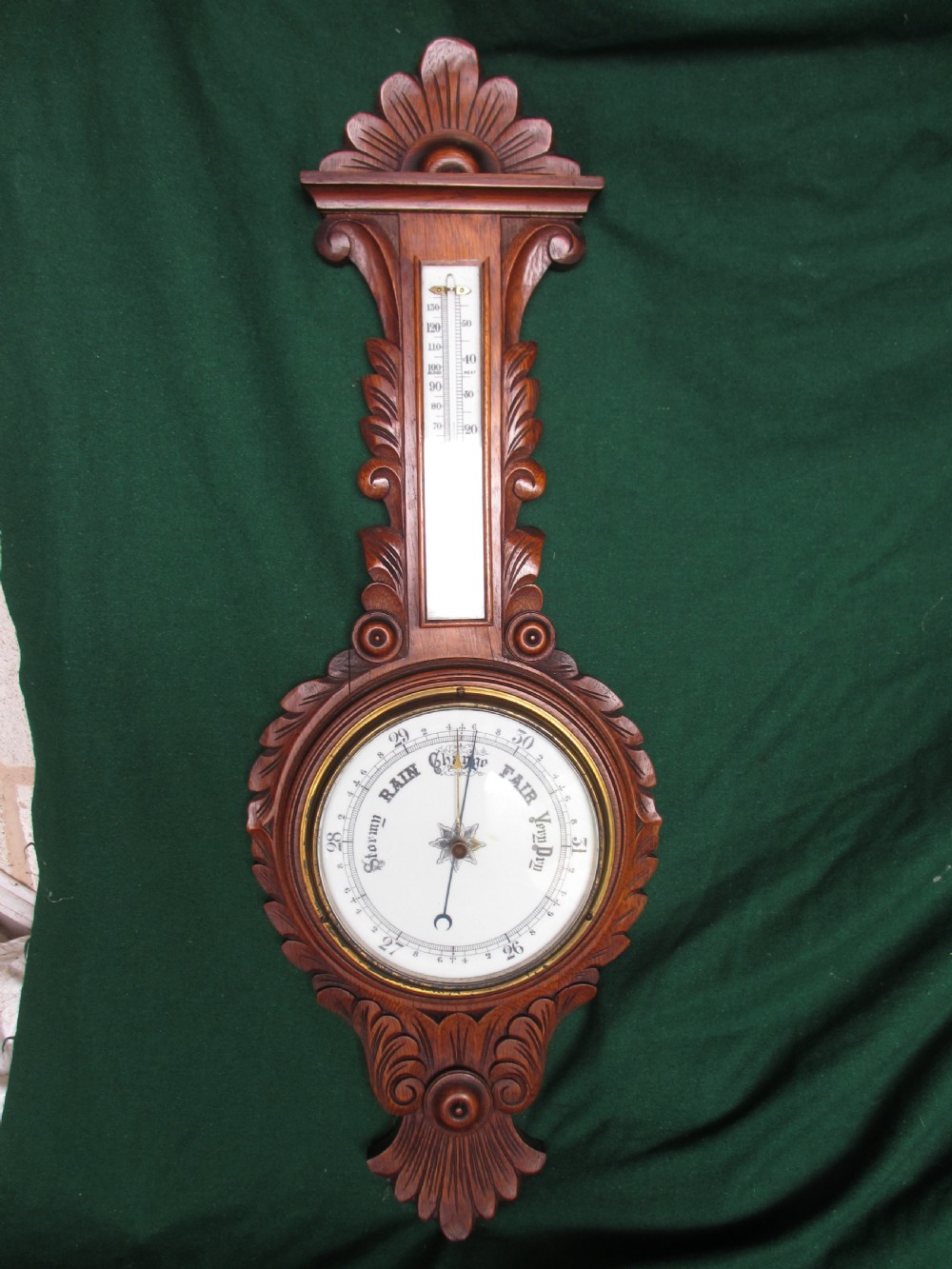 A Victorian carved oak wall barometer featuring an upper thermometer, intricate leaf scrollwork motifs, and a circular white dial with a brass bezel.