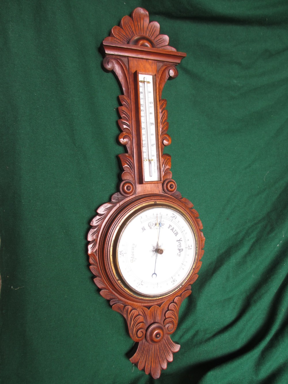 A Victorian carved oak wall barometer featuring an upper thermometer, intricate leaf scrollwork motifs, and a circular white dial with a brass bezel. View II