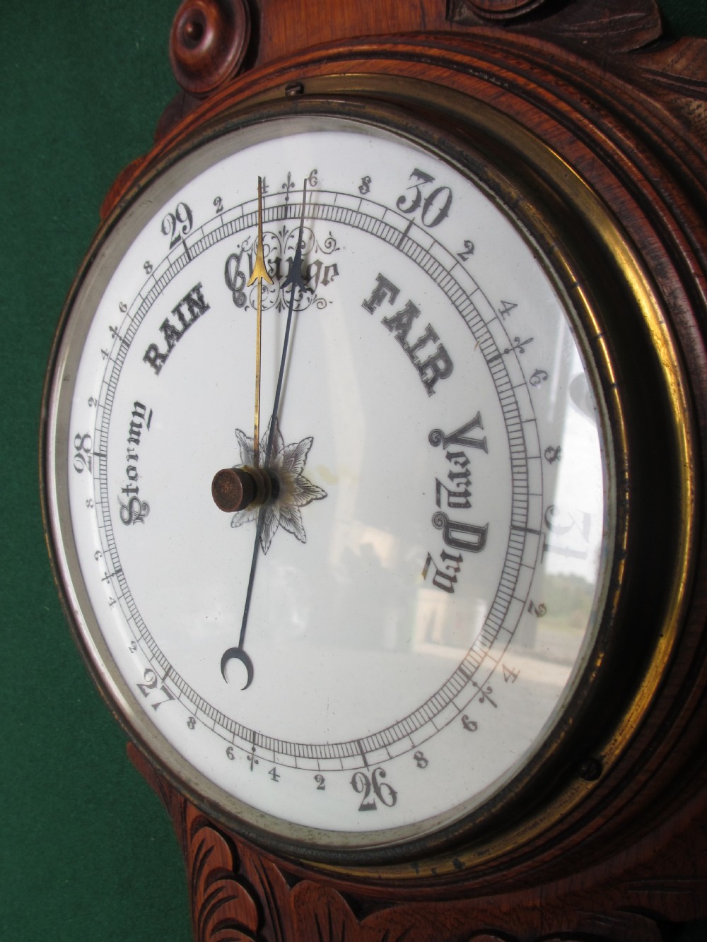 A Victorian carved oak wall barometer featuring an upper thermometer, intricate leaf scrollwork motifs, and a circular white dial with a brass bezel. View VII