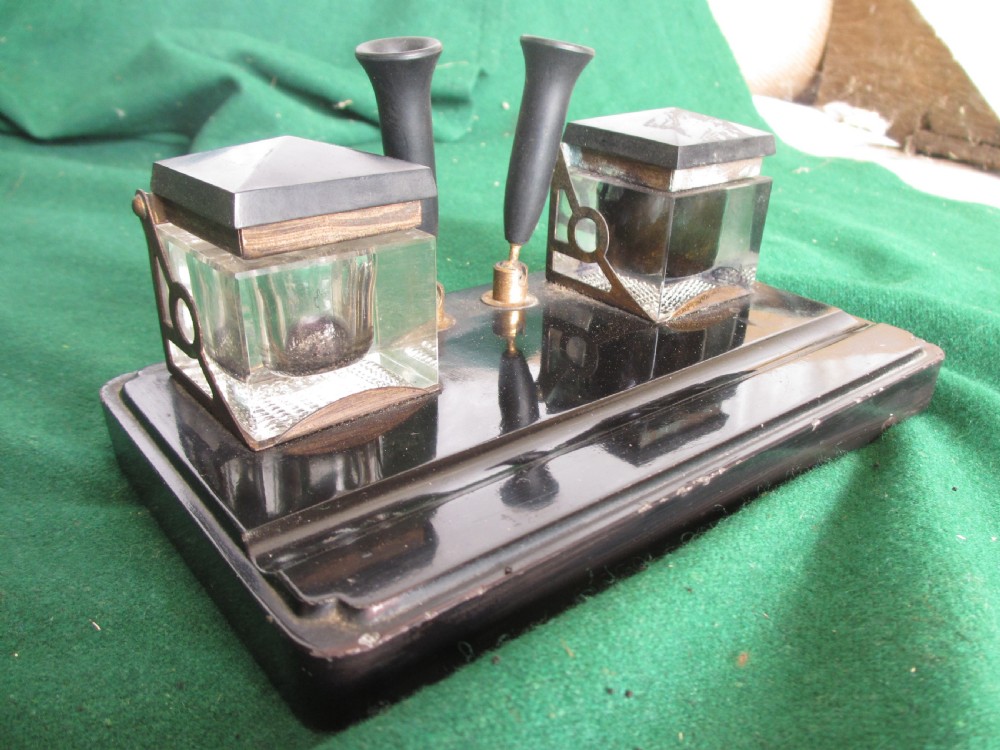 A Victorian inkstand featuring a black base with a front pen groove, two upright trumpet pen rests, and two square glass inkwells with dark lids.