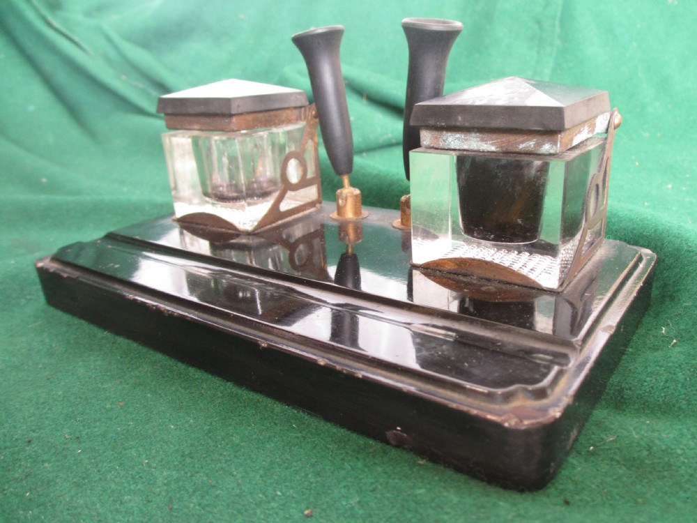 A Victorian inkstand featuring a black base with a front pen groove, two upright trumpet pen rests, and two square glass inkwells with dark lids. View IV