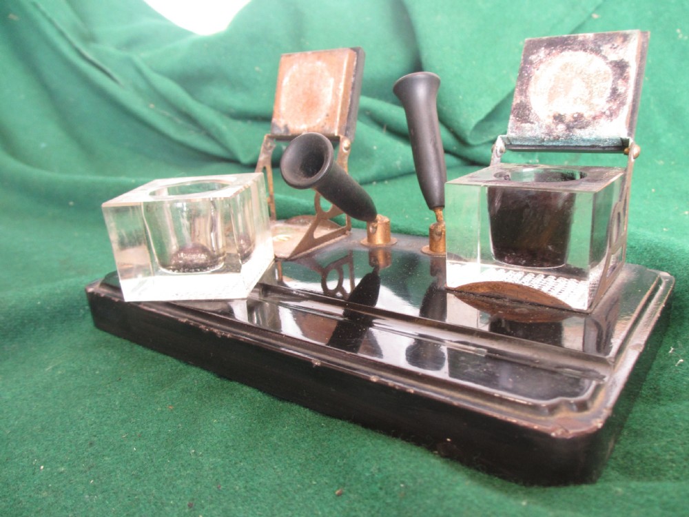 A Victorian inkstand featuring a black base with a front pen groove, two upright trumpet pen rests, and two square glass inkwells with dark lids. View V