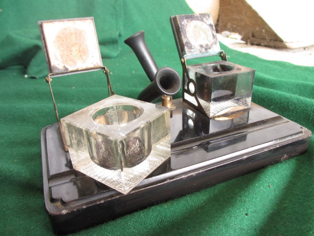 A Victorian inkstand featuring a black base with a front pen groove, two upright trumpet pen rests, and two square glass inkwells with dark lids. View VI
