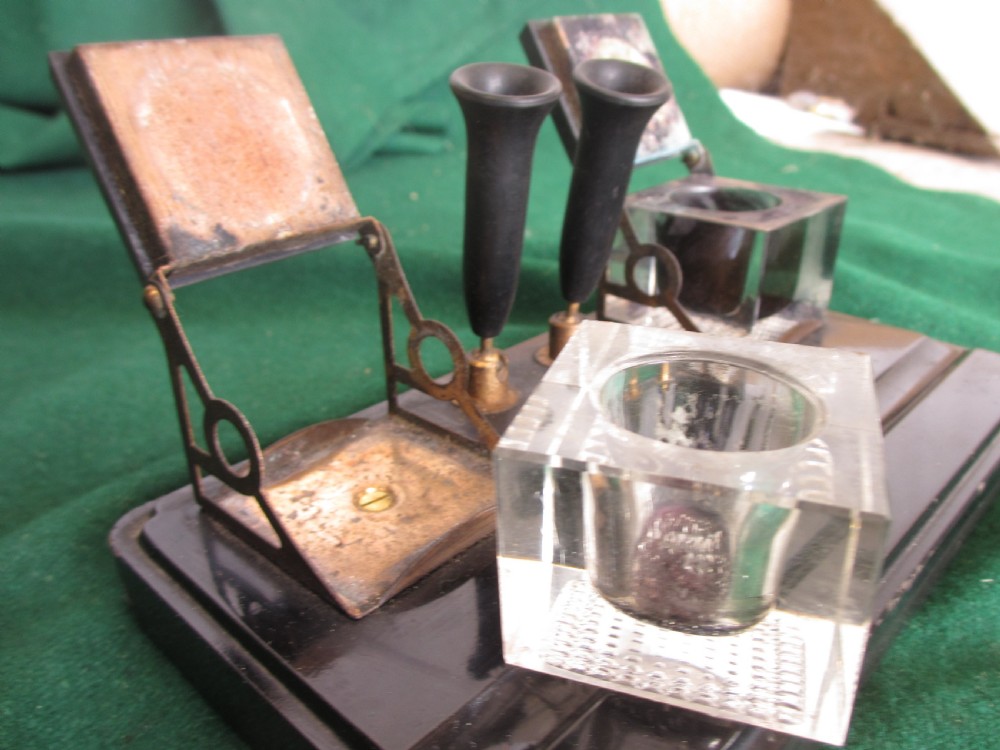 A Victorian inkstand featuring a black base with a front pen groove, two upright trumpet pen rests, and two square glass inkwells with dark lids. View IX