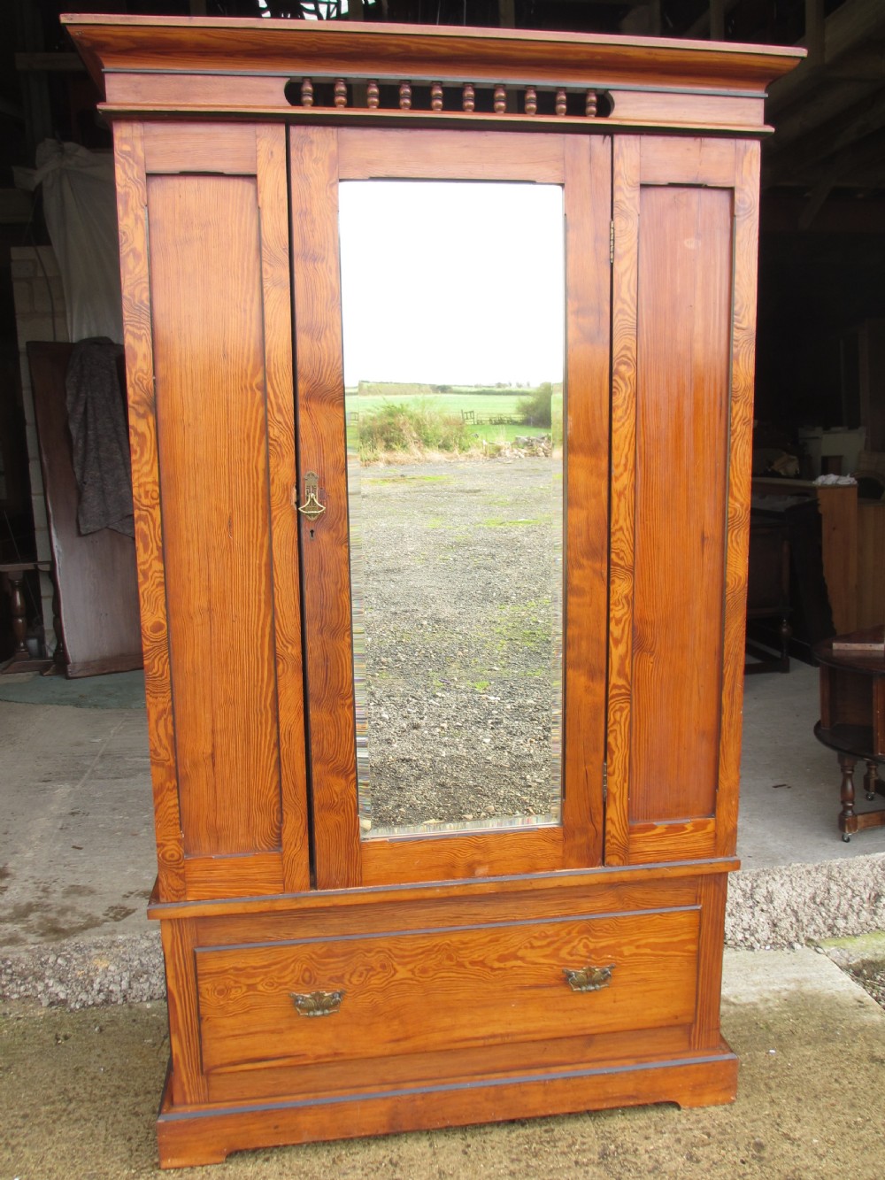 A Victorian pitch pine freestanding wardrobe featuring a central bevelled mirror door, upper spindle gallery, panelled sides, and a single lower drawer.