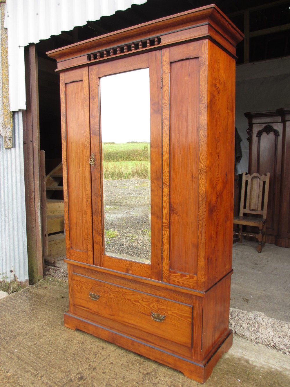 A Victorian pitch pine freestanding wardrobe featuring a central bevelled mirror door, upper spindle gallery, panelled sides, and a single lower drawer. View II