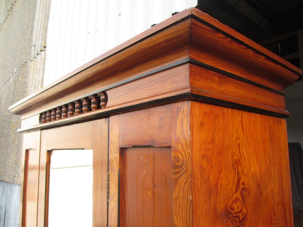 A Victorian pitch pine freestanding wardrobe featuring a central bevelled mirror door, upper spindle gallery, panelled sides, and a single lower drawer. View IV