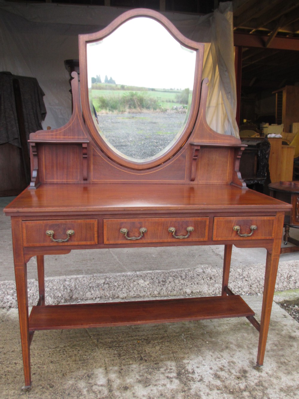 An Edwardian mahogany dressing table featuring a central shield-shaped swing mirror, crossbanded inlay details, three drawers with brass swan-neck handles, tapered legs, and a lower undertier shelf.