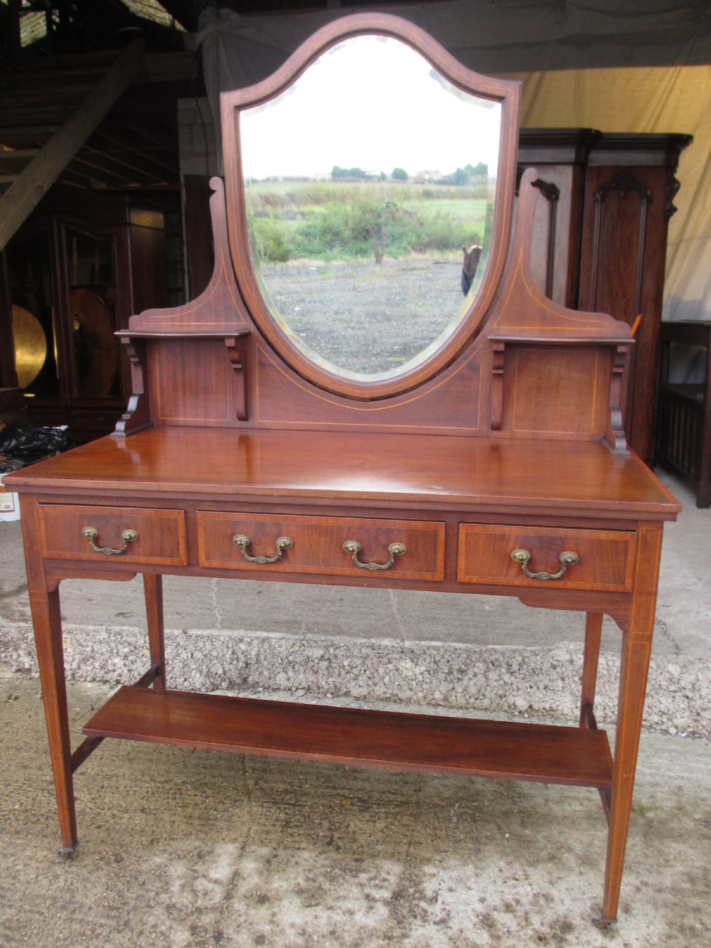 An Edwardian mahogany dressing table featuring a central shield-shaped swing mirror, crossbanded inlay details, three drawers with brass swan-neck handles, tapered legs, and a lower undertier shelf. View II