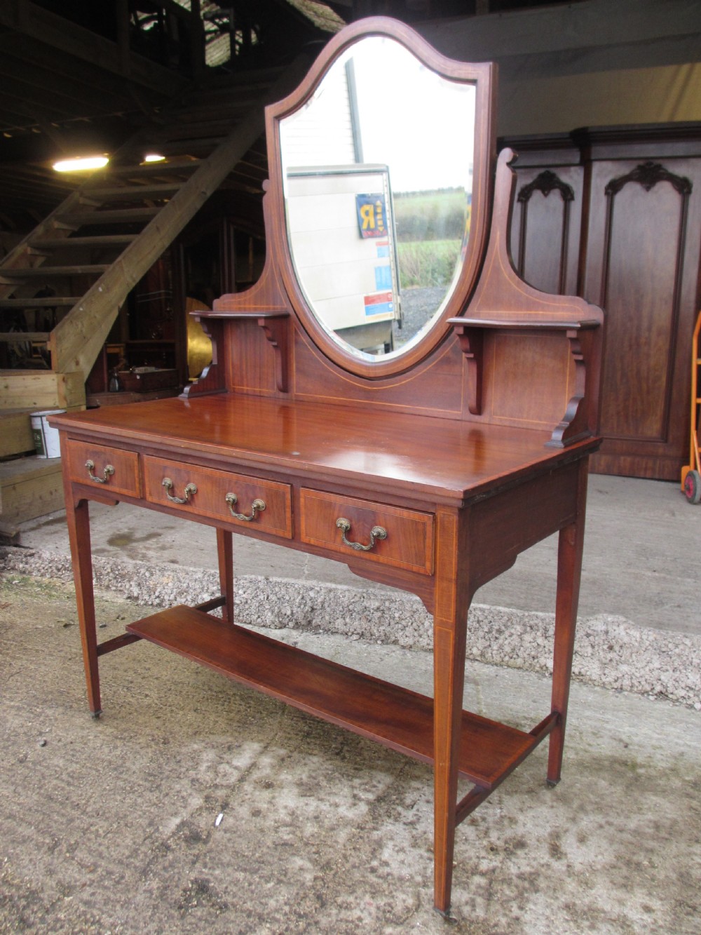 An Edwardian mahogany dressing table featuring a central shield-shaped swing mirror, crossbanded inlay details, three drawers with brass swan-neck handles, tapered legs, and a lower undertier shelf. View III