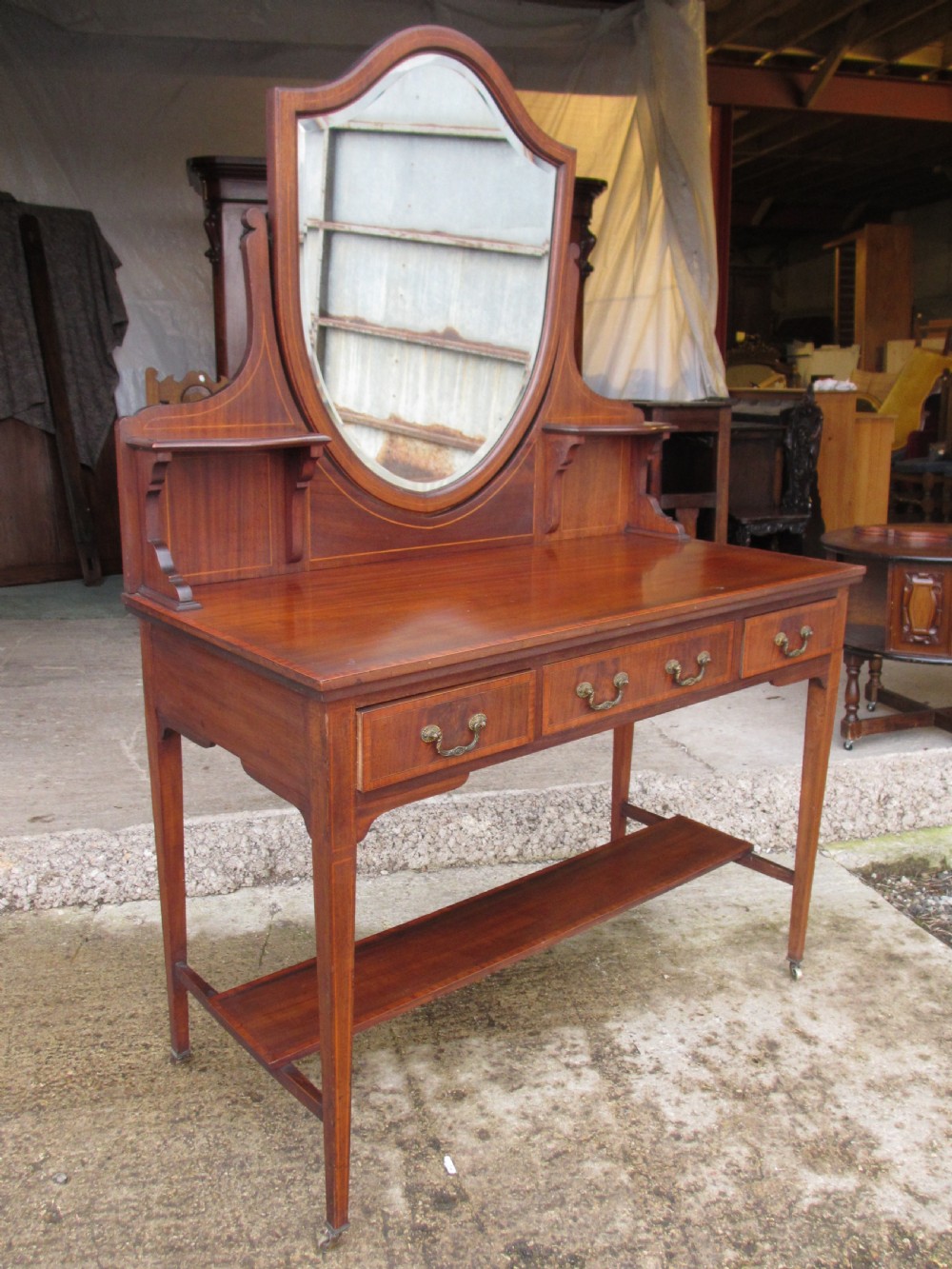 An Edwardian mahogany dressing table featuring a central shield-shaped swing mirror, crossbanded inlay details, three drawers with brass swan-neck handles, tapered legs, and a lower undertier shelf. View IV