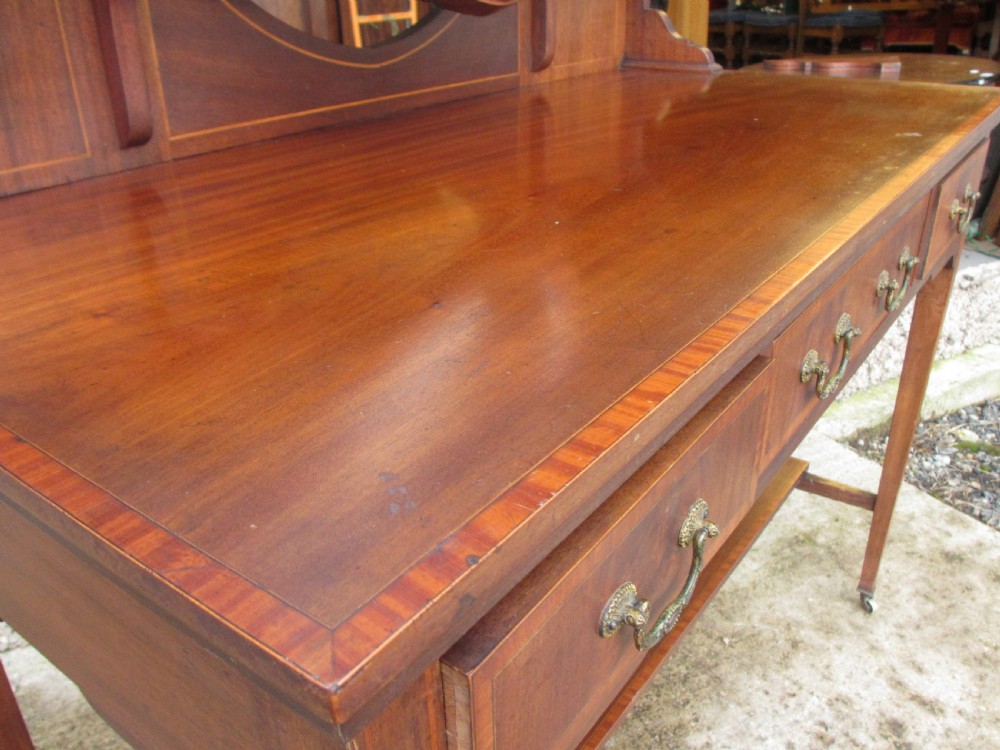 An Edwardian mahogany dressing table featuring a central shield-shaped swing mirror, crossbanded inlay details, three drawers with brass swan-neck handles, tapered legs, and a lower undertier shelf. View VI