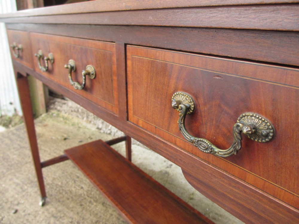 An Edwardian mahogany dressing table featuring a central shield-shaped swing mirror, crossbanded inlay details, three drawers with brass swan-neck handles, tapered legs, and a lower undertier shelf. View XI