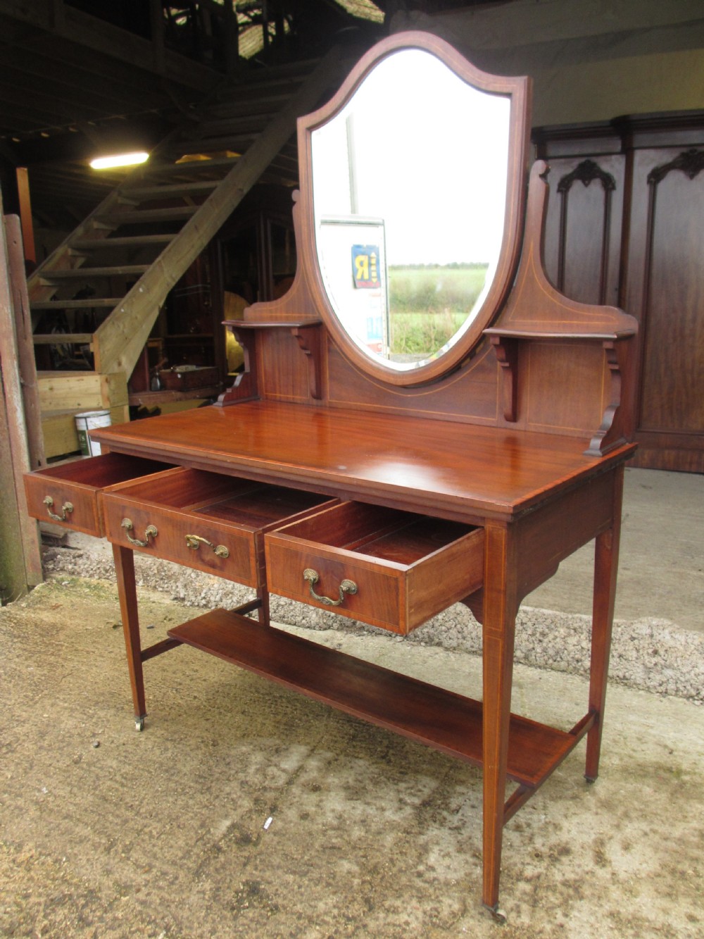 An Edwardian mahogany dressing table featuring a central shield-shaped swing mirror, crossbanded inlay details, three drawers with brass swan-neck handles, tapered legs, and a lower undertier shelf. View XII