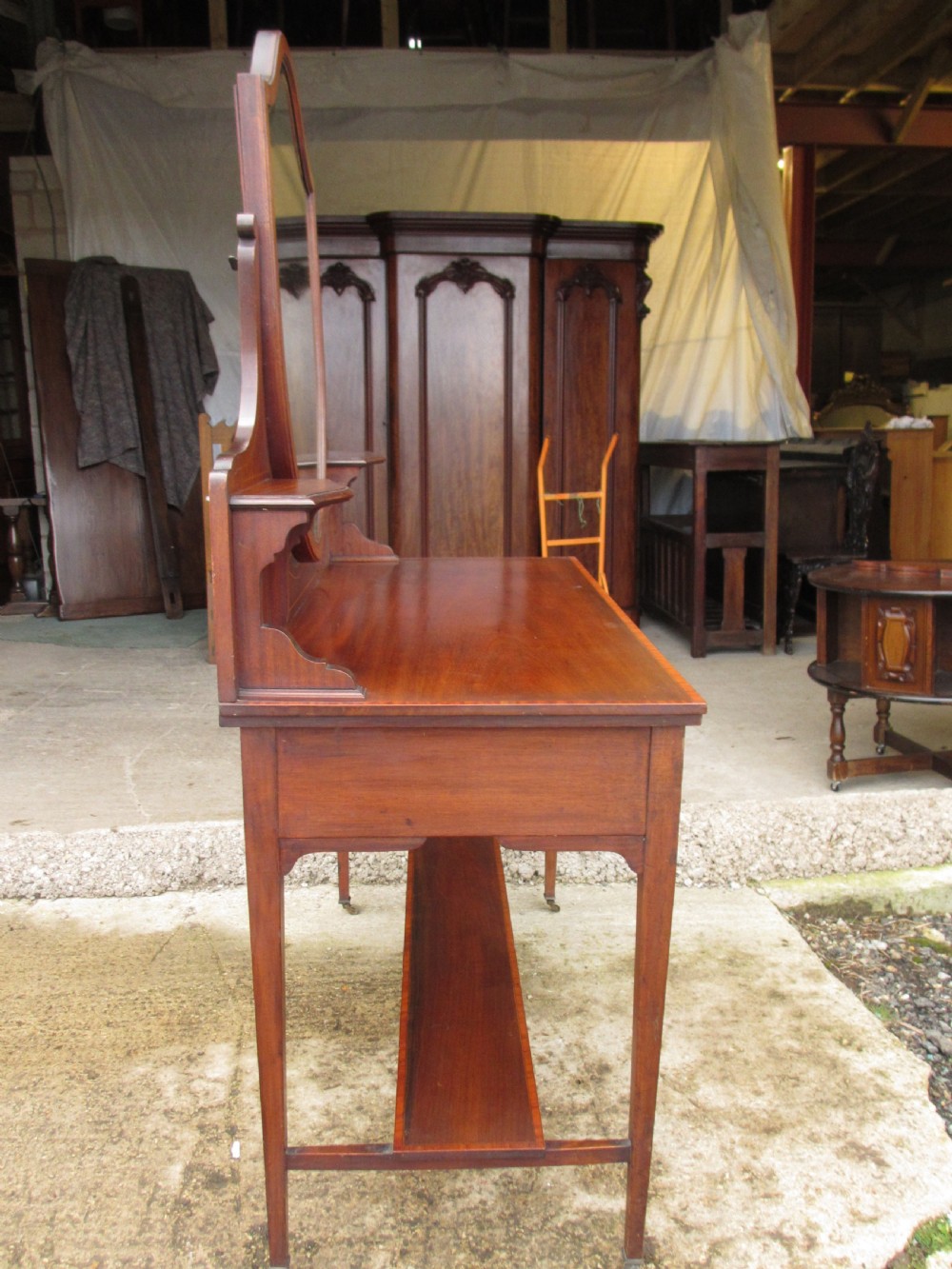 An Edwardian mahogany dressing table featuring a central shield-shaped swing mirror, crossbanded inlay details, three drawers with brass swan-neck handles, tapered legs, and a lower undertier shelf. View XIII