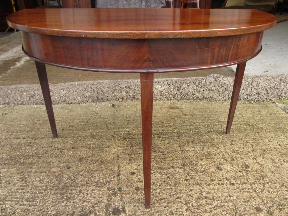 A 19th-century mahogany demi-lune console table featuring a semi-circular polished surface, a deep apron, and three slender tapered legs.