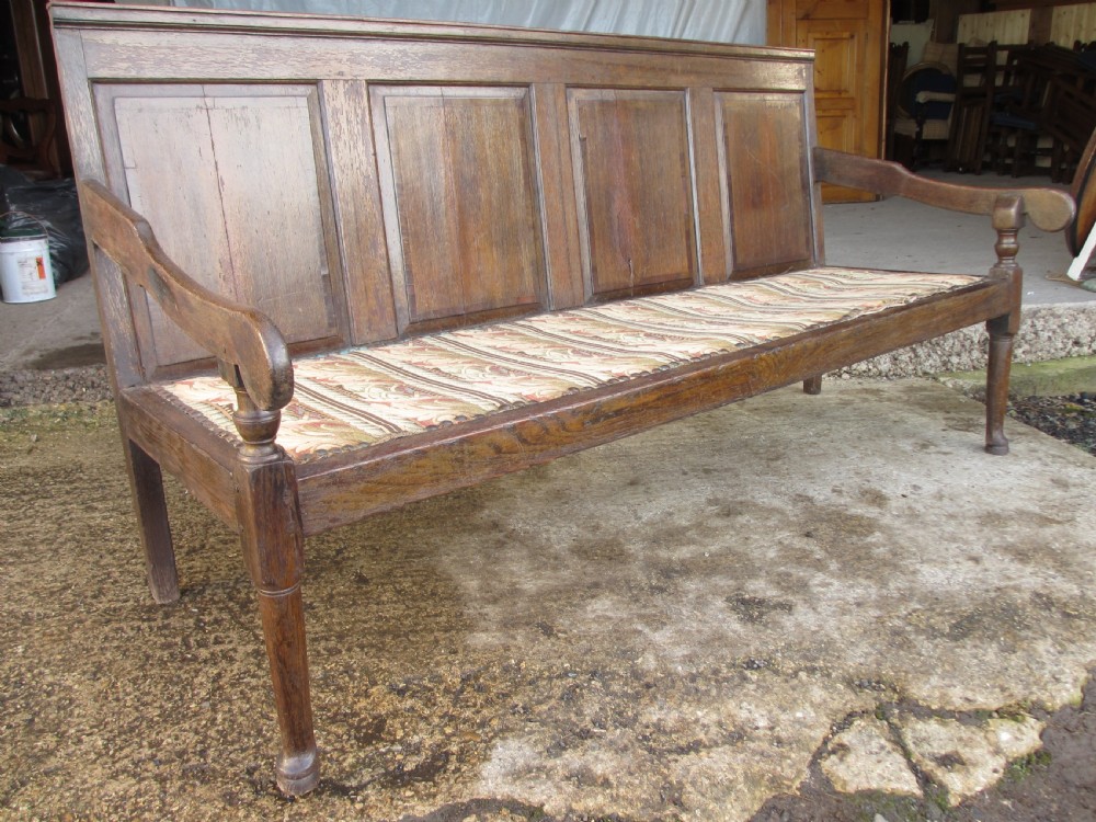 An 18th-century oak settle featuring a four-panel wooden backrest, turned front legs, shaped armrests, and an upholstered patterned seat cushion.