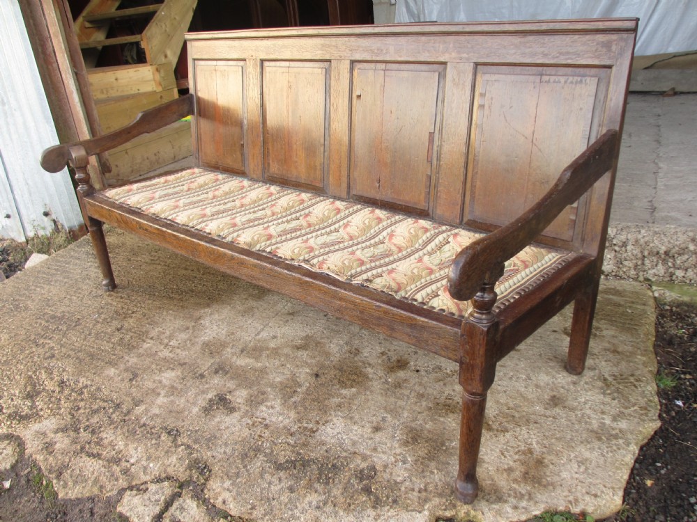 An 18th-century oak settle featuring a four-panel wooden backrest, turned front legs, shaped armrests, and an upholstered patterned seat cushion. View II