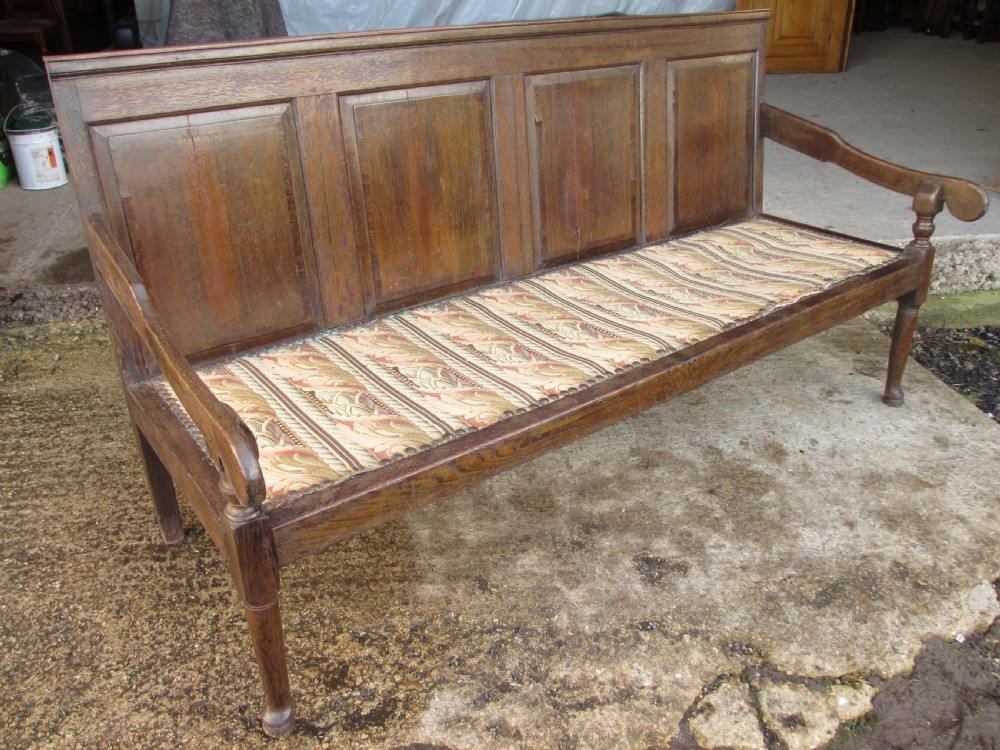 An 18th-century oak settle featuring a four-panel wooden backrest, turned front legs, shaped armrests, and an upholstered patterned seat cushion. View IV
