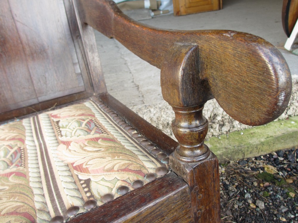 An 18th-century oak settle featuring a four-panel wooden backrest, turned front legs, shaped armrests, and an upholstered patterned seat cushion. View VII