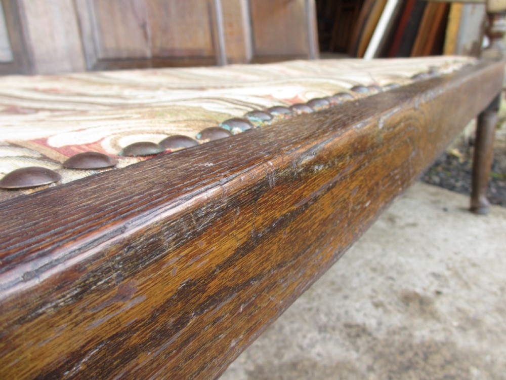 An 18th-century oak settle featuring a four-panel wooden backrest, turned front legs, shaped armrests, and an upholstered patterned seat cushion. View X
