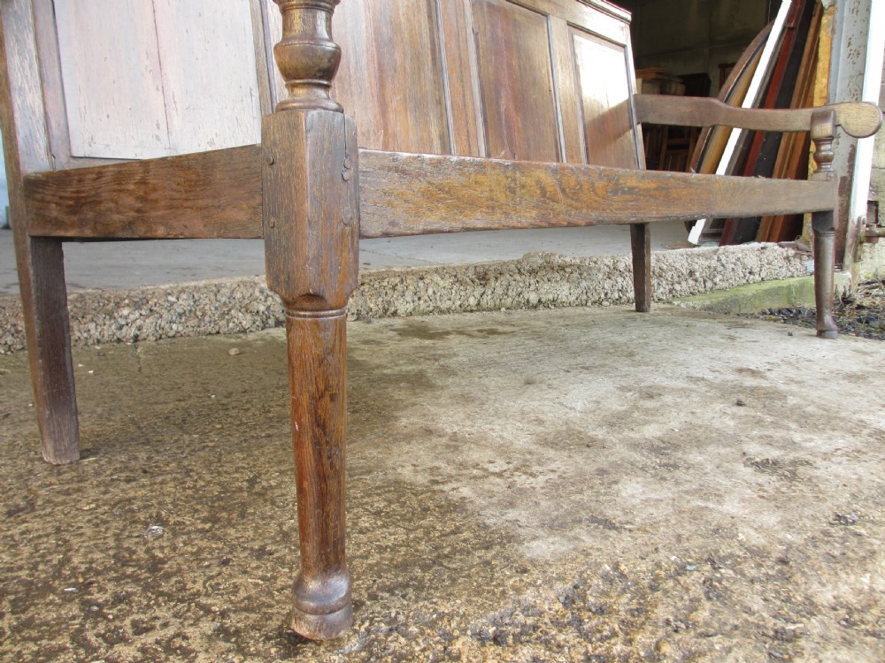 An 18th-century oak settle featuring a four-panel wooden backrest, turned front legs, shaped armrests, and an upholstered patterned seat cushion. View XI