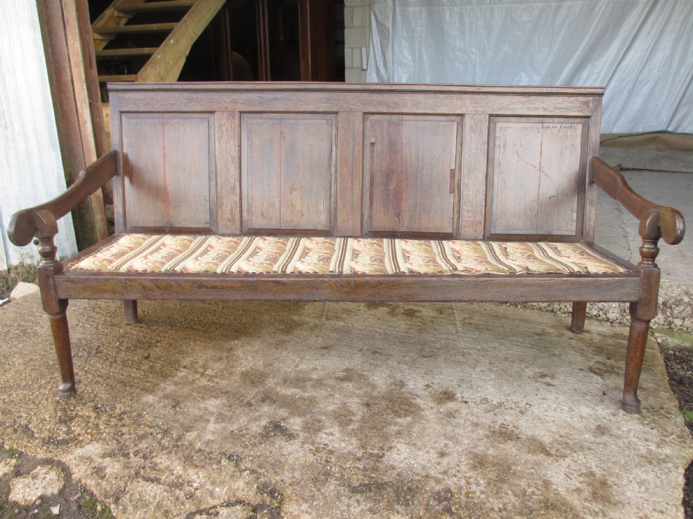 An 18th-century oak settle featuring a four-panel wooden backrest, turned front legs, shaped armrests, and an upholstered patterned seat cushion. View XIV