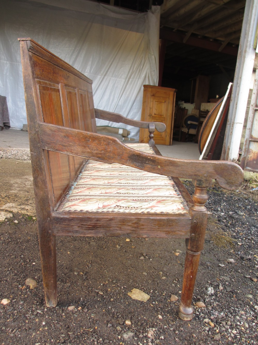 An 18th-century oak settle featuring a four-panel wooden backrest, turned front legs, shaped armrests, and an upholstered patterned seat cushion. View XV