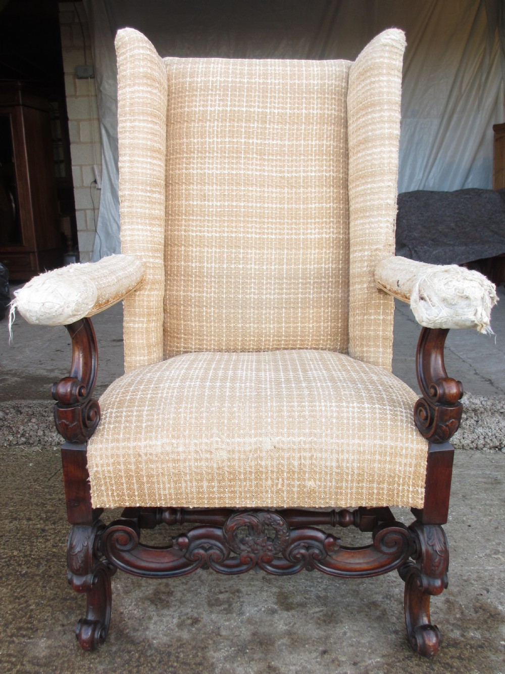 A 19th-century high wingback armchair featuring an intricately carved walnut frame with scroll armrests and neutral woven fabric upholstery.