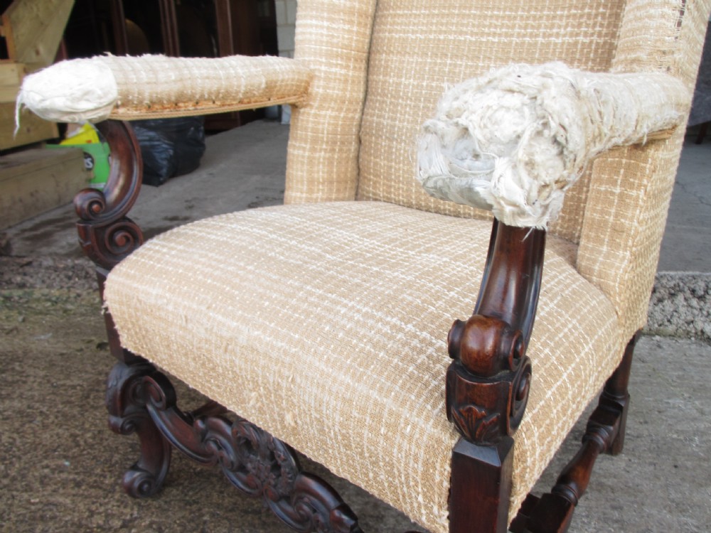 A 19th-century high wingback armchair featuring an intricately carved walnut frame with scroll armrests and neutral woven fabric upholstery. View V