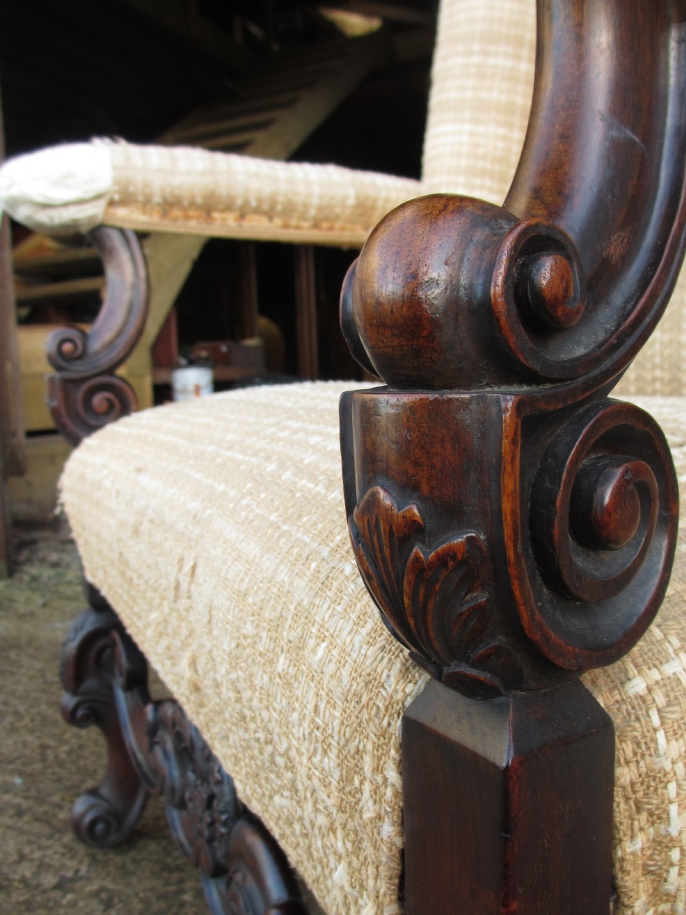 A 19th-century high wingback armchair featuring an intricately carved walnut frame with scroll armrests and neutral woven fabric upholstery. View X