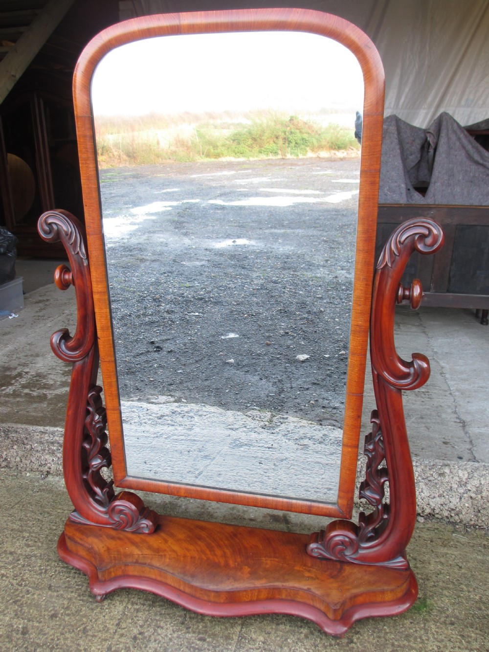 A 19th-century flame mahogany cheval mirror featuring an arched tilting frame suspended between carved scroll supports on a serpentine base.