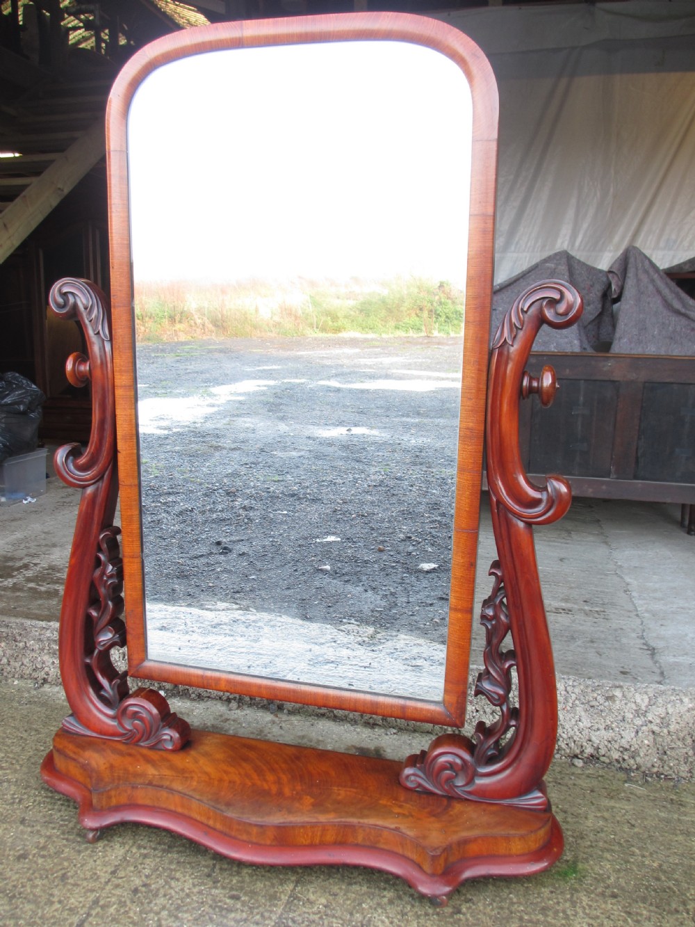 A 19th-century flame mahogany cheval mirror featuring an arched tilting frame suspended between carved scroll supports on a serpentine base. View II