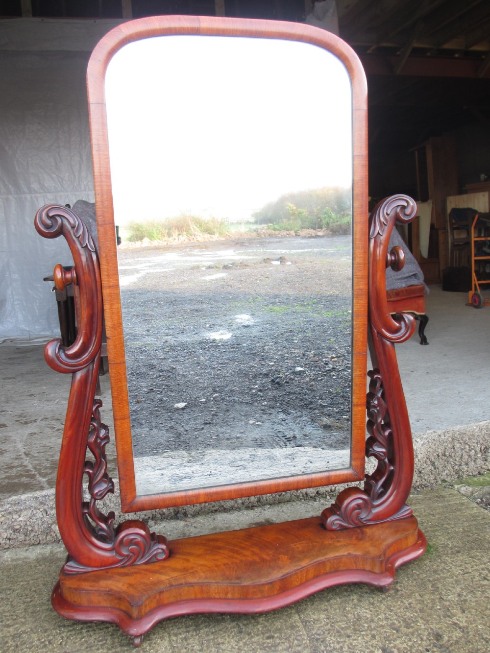 A 19th-century flame mahogany cheval mirror featuring an arched tilting frame suspended between carved scroll supports on a serpentine base. View III