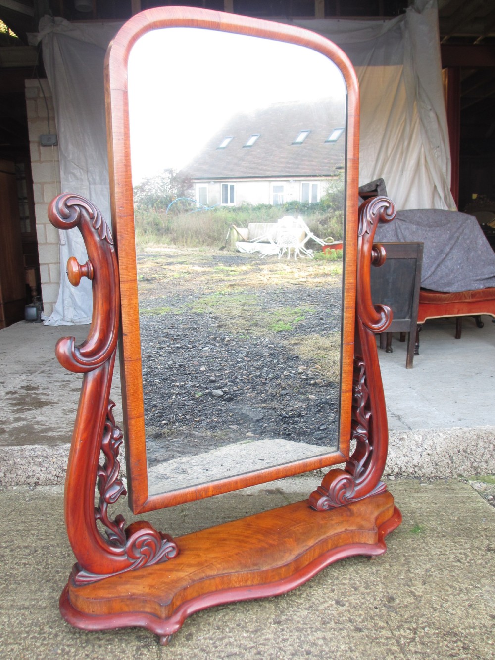 A 19th-century flame mahogany cheval mirror featuring an arched tilting frame suspended between carved scroll supports on a serpentine base. View IV