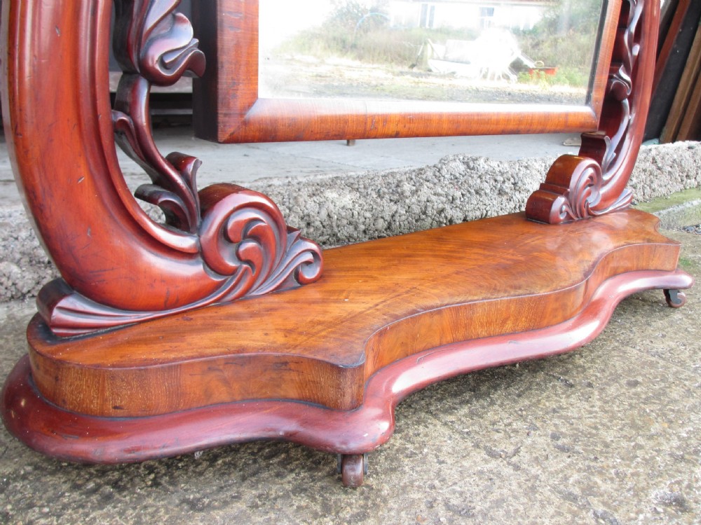 A 19th-century flame mahogany cheval mirror featuring an arched tilting frame suspended between carved scroll supports on a serpentine base. View VIII