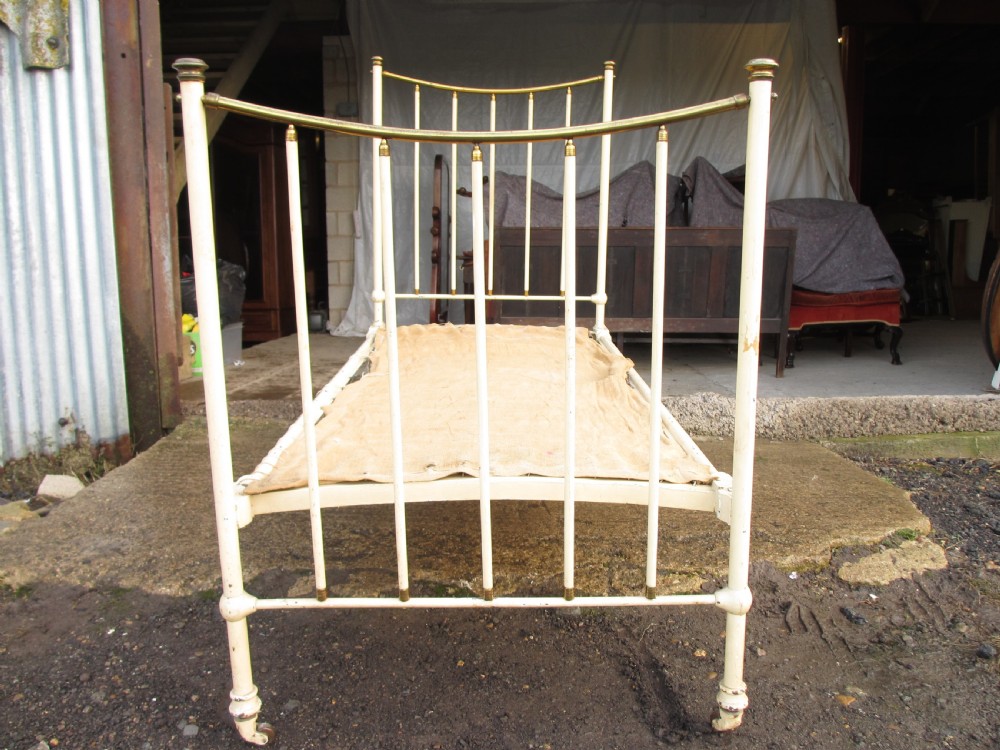 An antique Victorian single bedstead featuring a painted off-white iron frame, brass top rails, vertical spindles, and castors.