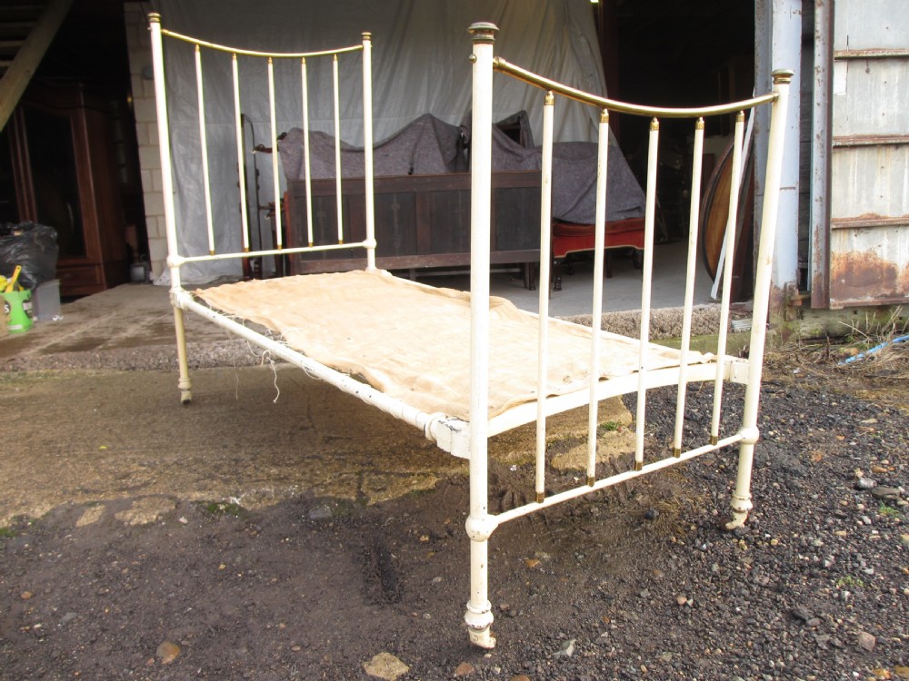 An antique Victorian single bedstead featuring a painted off-white iron frame, brass top rails, vertical spindles, and castors. View II