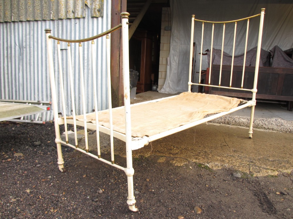 An antique Victorian single bedstead featuring a painted off-white iron frame, brass top rails, vertical spindles, and castors. View III