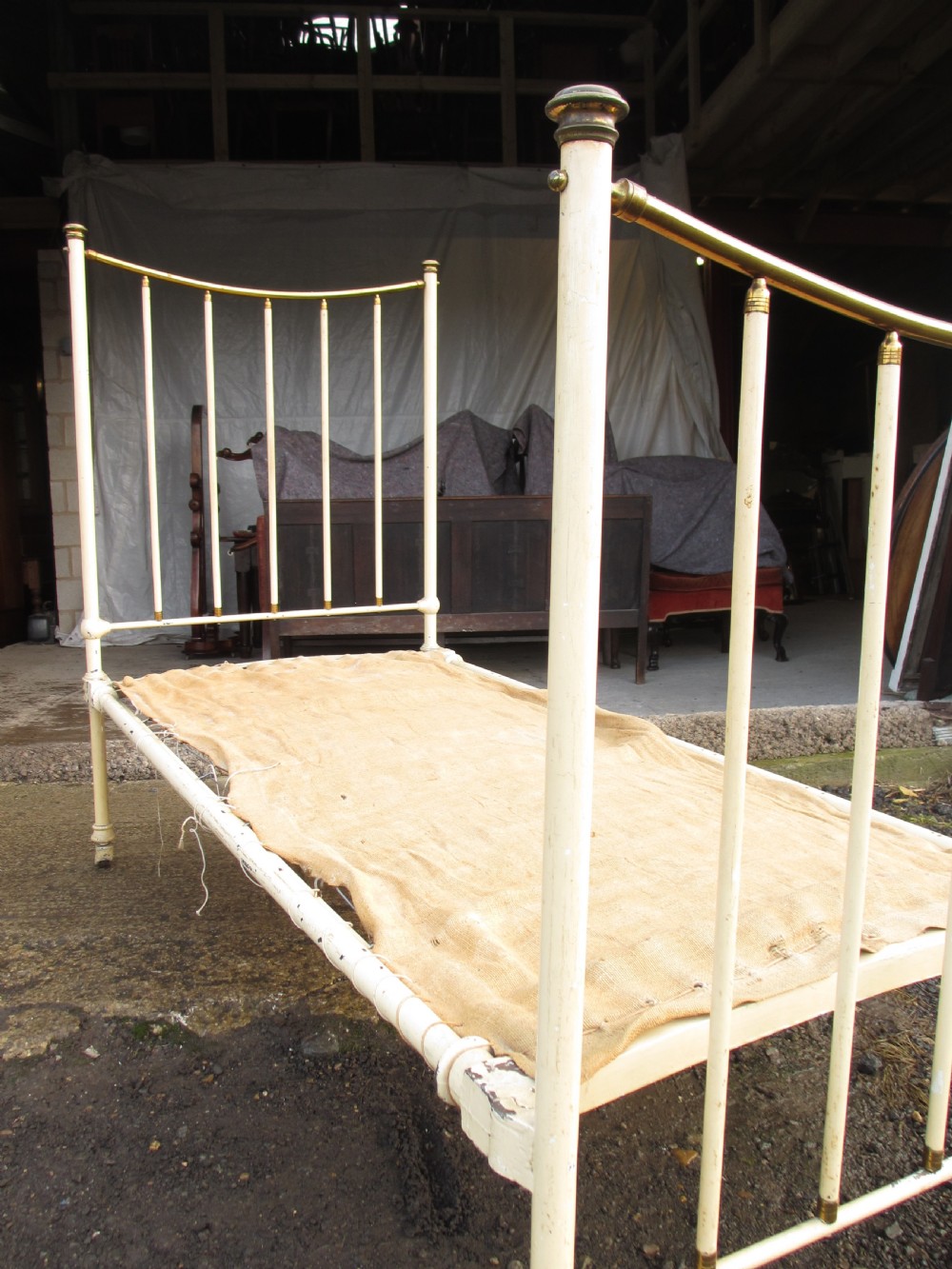 An antique Victorian single bedstead featuring a painted off-white iron frame, brass top rails, vertical spindles, and castors. View IV