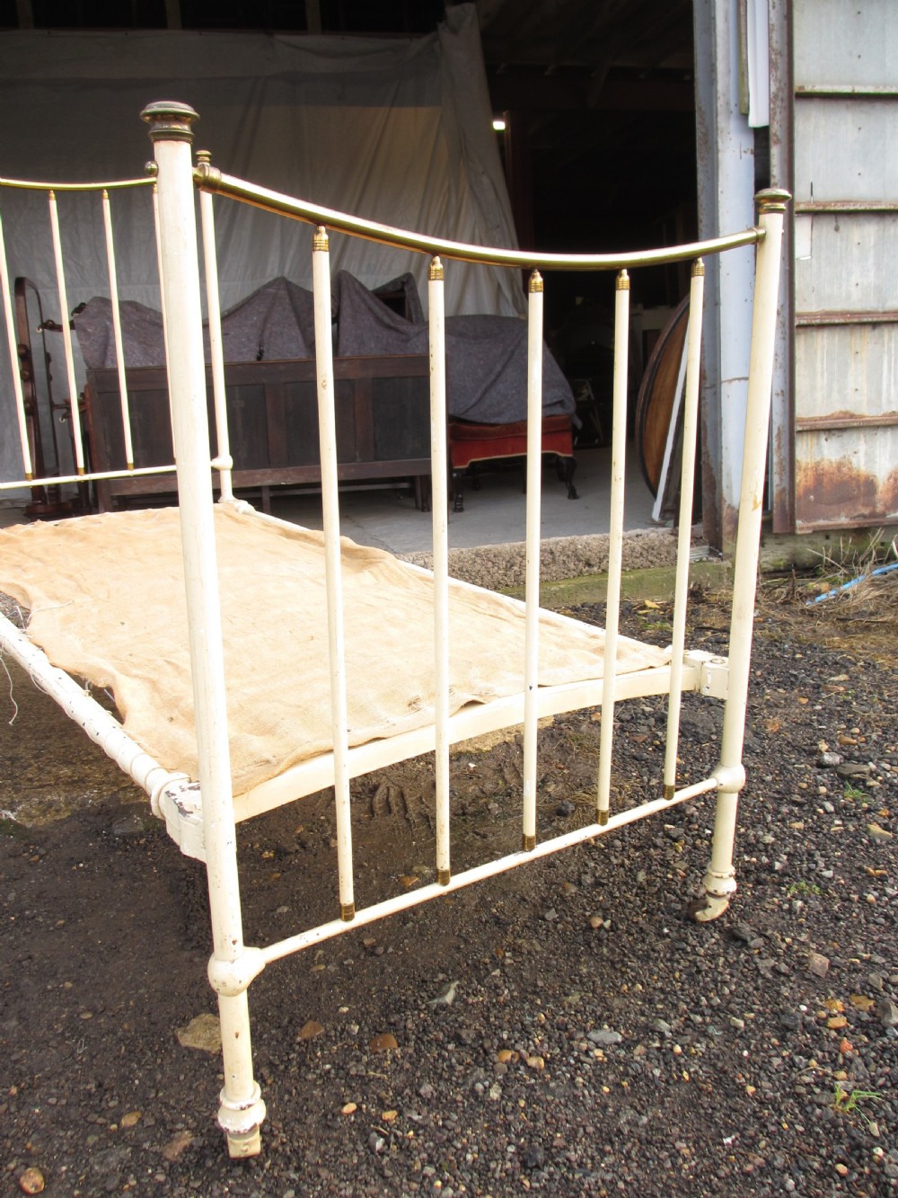 An antique Victorian single bedstead featuring a painted off-white iron frame, brass top rails, vertical spindles, and castors. View V