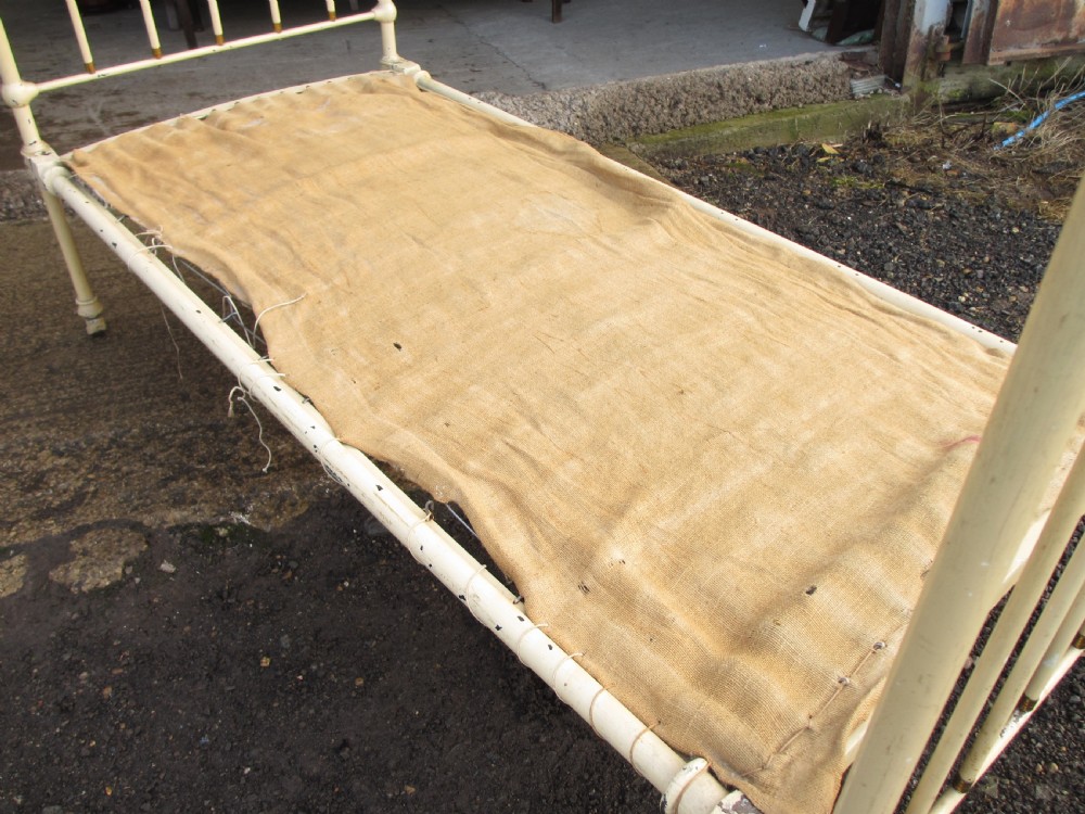 An antique Victorian single bedstead featuring a painted off-white iron frame, brass top rails, vertical spindles, and castors. View VII