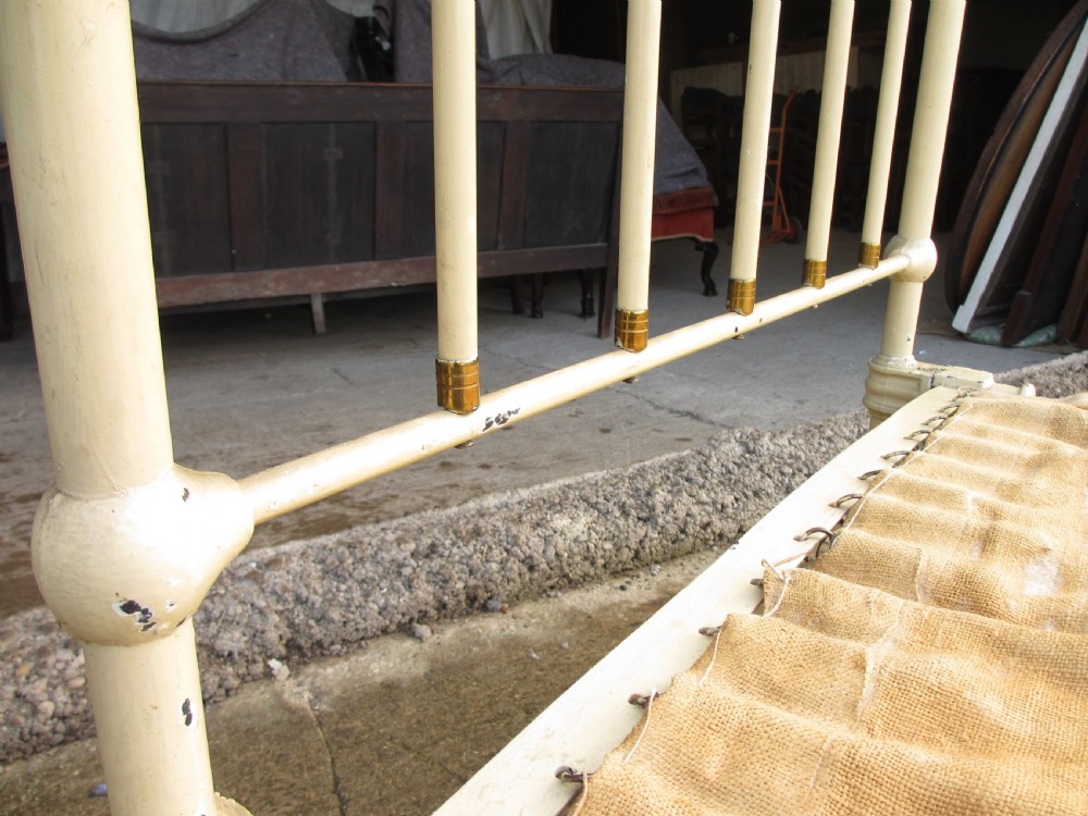 An antique Victorian single bedstead featuring a painted off-white iron frame, brass top rails, vertical spindles, and castors. View IX