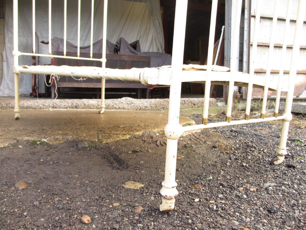 An antique Victorian single bedstead featuring a painted off-white iron frame, brass top rails, vertical spindles, and castors. View XII