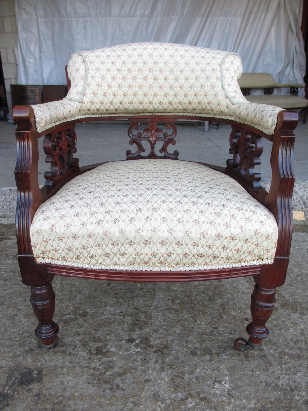 A pair of antique mahogany tub armchairs featuring cream patterned fabric upholstery, openwork carved back splats, and turned front legs on castors. View IV